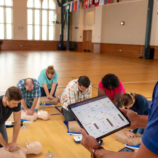 A group of students practicing CPR with Adult Prestan Professional Series 2000 Manikins with an instructor monitoring progress on a tablet.