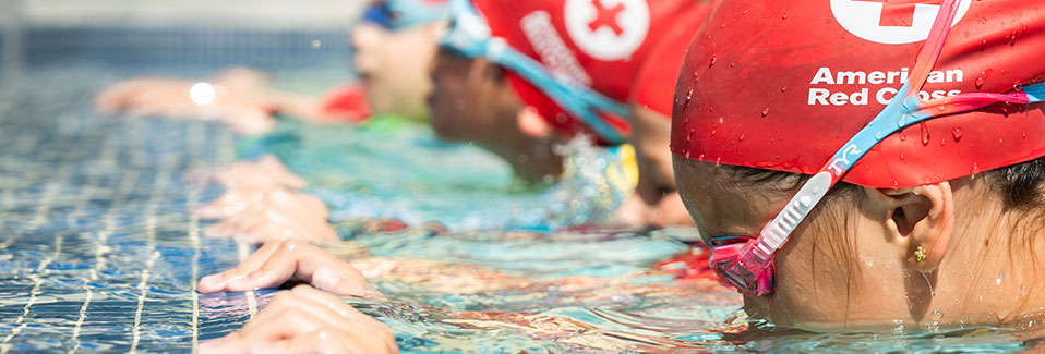 A group of swimming students in a pool wearing goggles and American Red Cross swim caps.