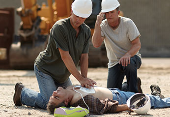 Two construction workers using an AED and performing CPR on a coworker in distress on the worksite.